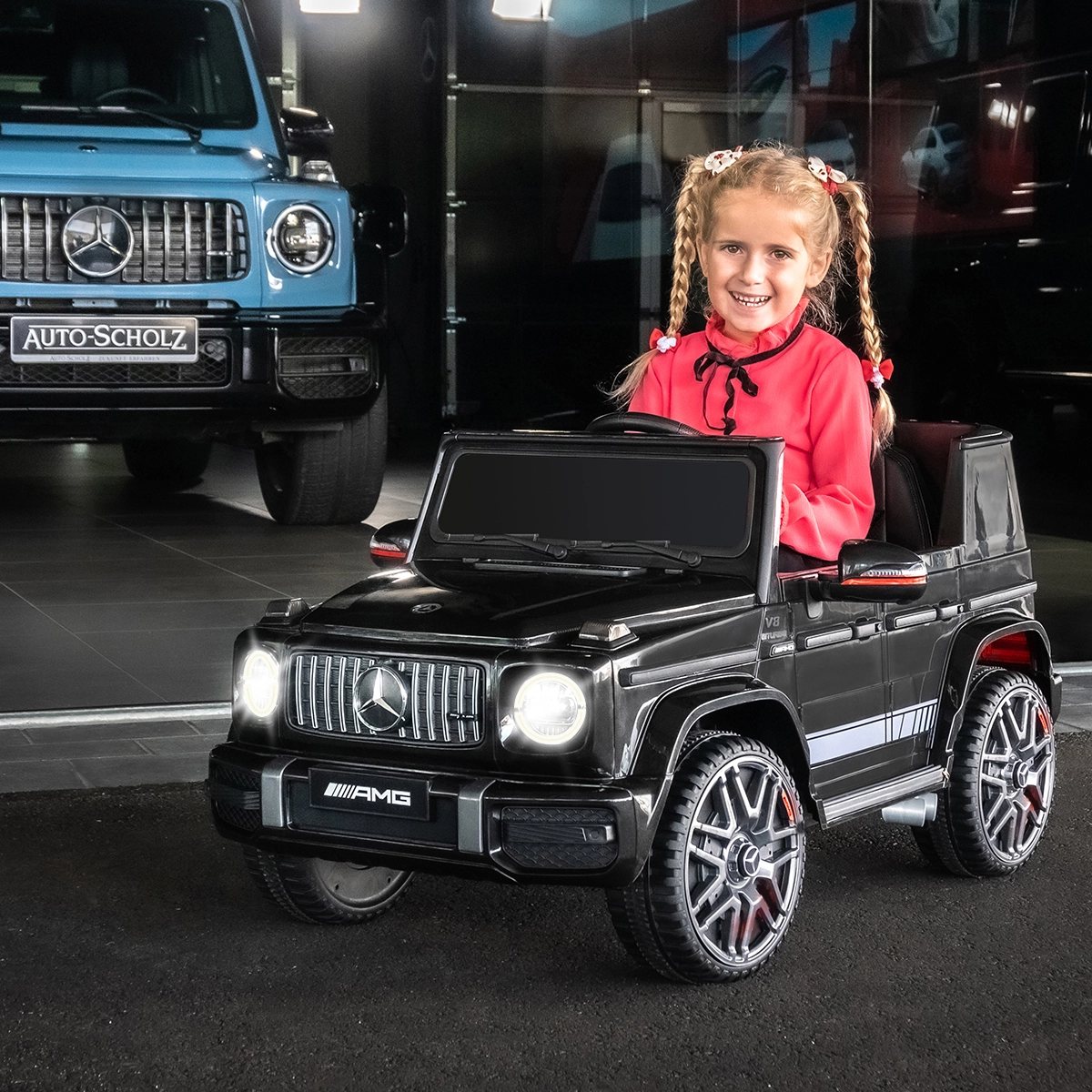 Strahlend helle LED-Scheinwerfer Mädchen sitzt lächelnd im Kinder-Elektroauto Mercedes AMG G63 W463